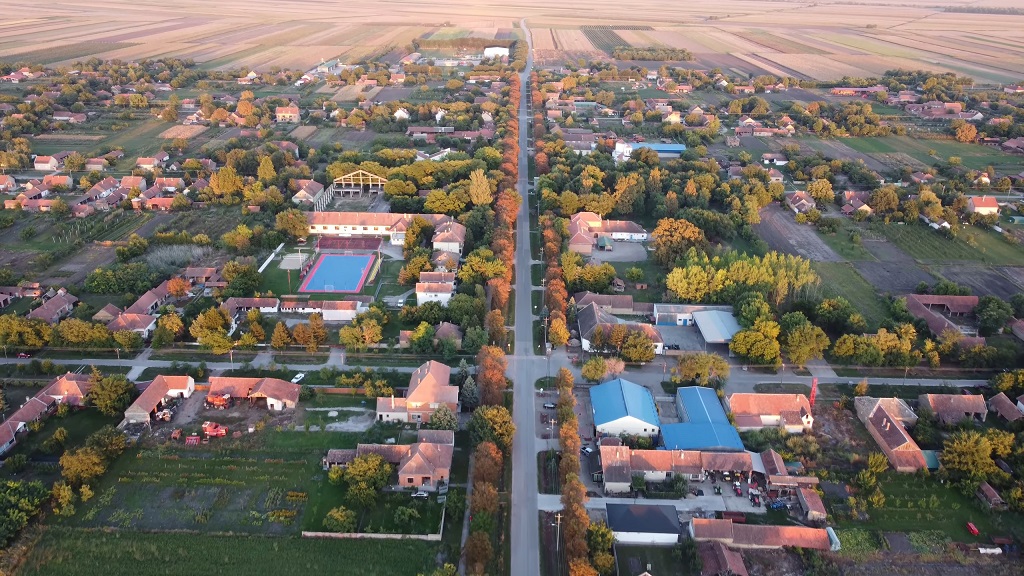 Panorama sela snimljena iz vazduha, sa urednim nizovima kuća, okućnicama i prostranim poljima koja se protežu do horizonta