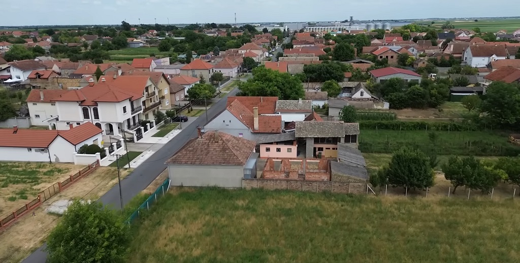 Pogled iz vazduha na naselje sa kućama i baštama u manjoj sredini, okruženo zelenim površinama